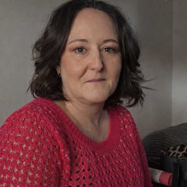Indoor portrait of Vicky Fowles sitting against a neutral wall. She has short dark brown hair and is wearing a bright pink knitted jumper. She is looking directly at the camera with a neutral expression
