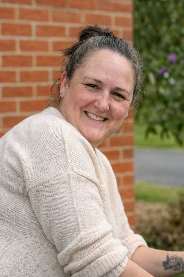 Outdoor photo of Vicky Fowles standing beside a brick wall. She is wearing a light beige knitted sweater and has her hair tied back. A tattoo is visible on her forearm. The background includes greenery, a road, and part of a garden area.