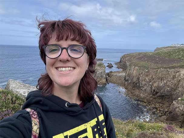 Ellen Gibson taking a selfie near a coastal cliff, wearing glasses and a black hoodie. The background shows rocky cliffs, grass, and the ocean.